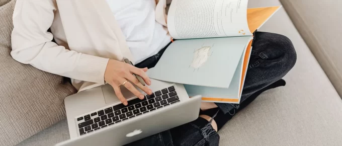 Person sitting on a sofa reading printed pages beside an open laptop