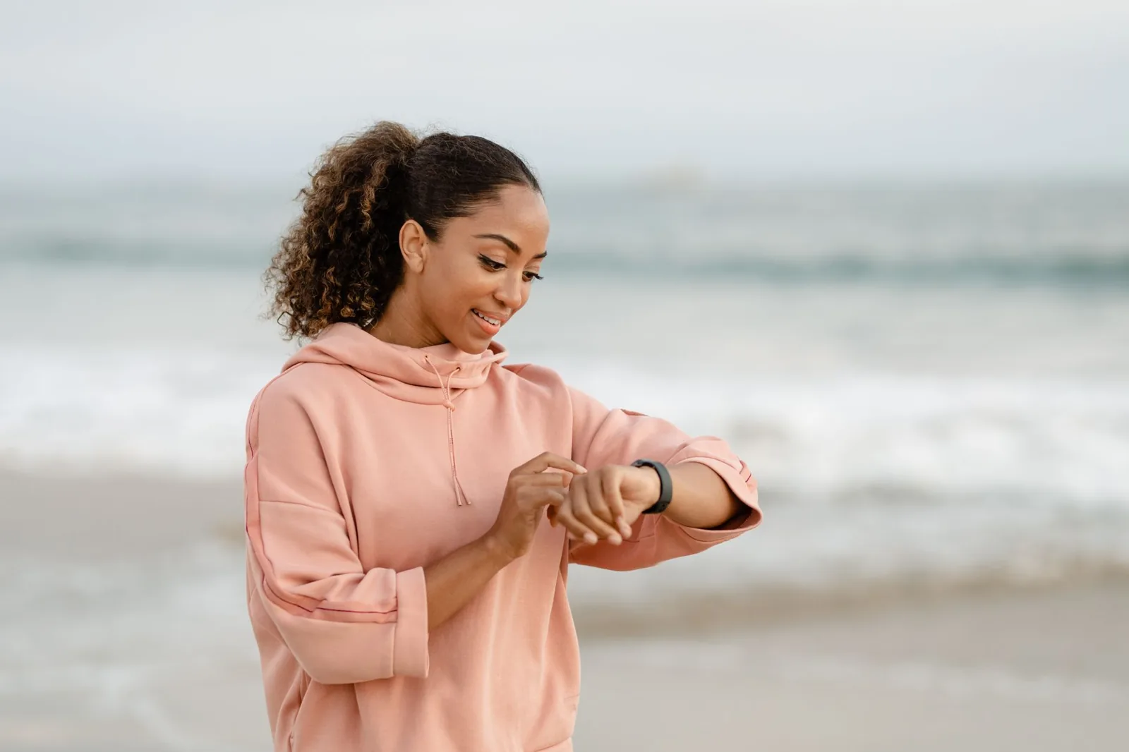 Woman in a pink hoodie checking her smartwatch on a beach